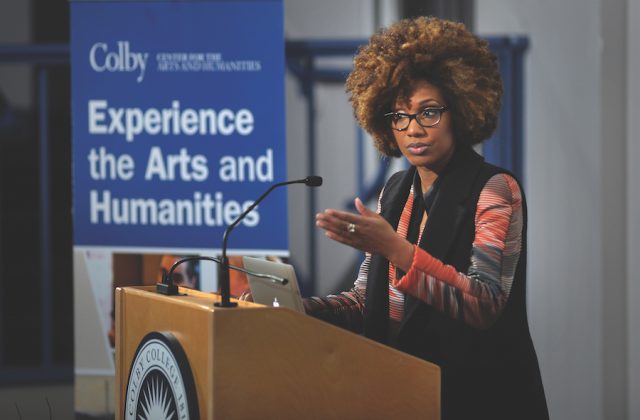 A speaker with curly hair presents at a podium with a banner behind reading Experience the Arts and Humanities