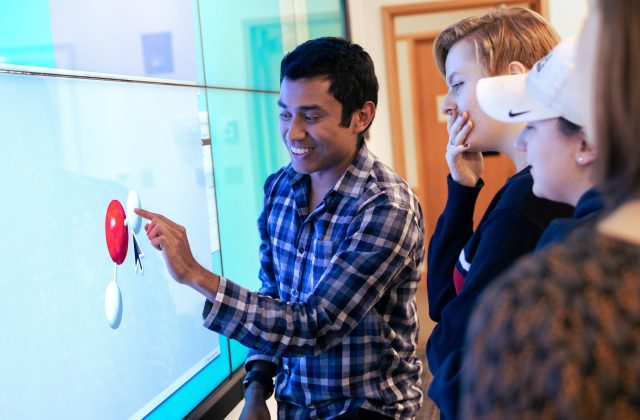 A man demonstrates an interactive display to a group of engaged students exploring molecular structures in a classroom