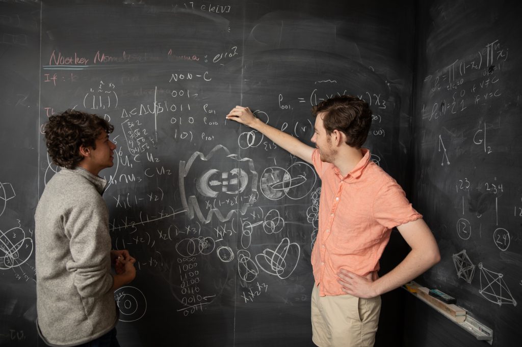 Two individuals discussing equations and diagrams on a chalkboard filled with mathematical notations and illustrations