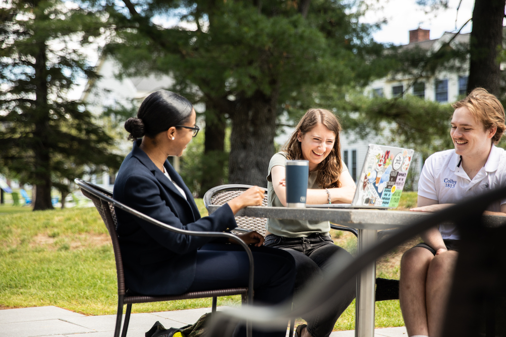 Three students engaged in conversation at an outdoor table surrounded by trees and a relaxed campus atmosphere