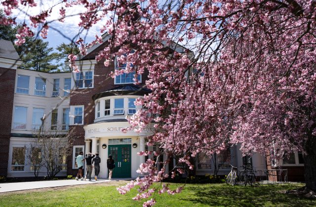 A red-brick and white-trimmed building partially framed by pink cherry blossoms in full bloom. A group of students walks toward the entrance.