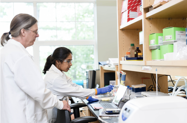 One older woman and one younger woman wearing white coats examine a sample in a lab.